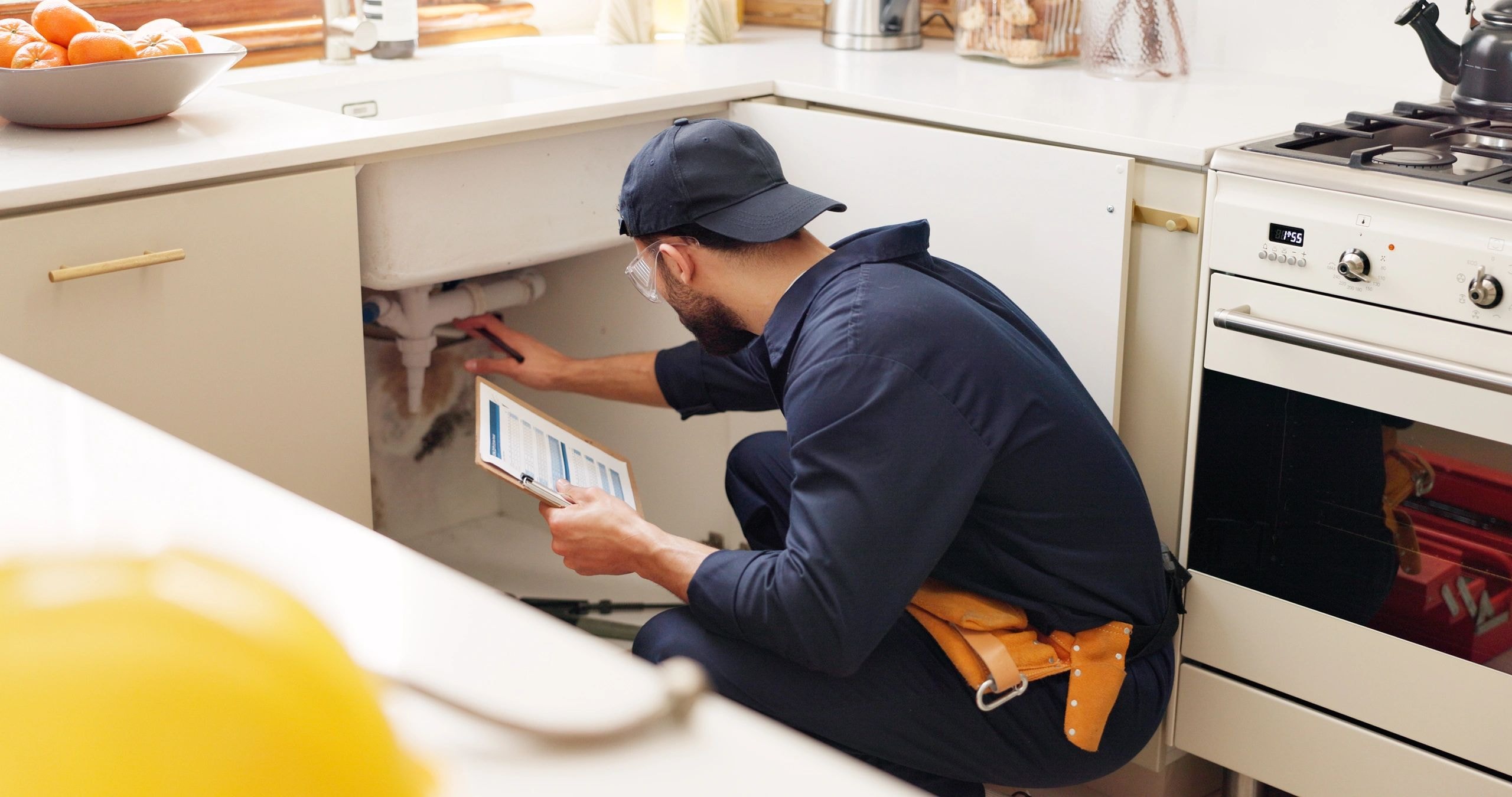 A plumber in navy work clothes inspecting the plumbing beneath a kitchen sink while holding a clipboard